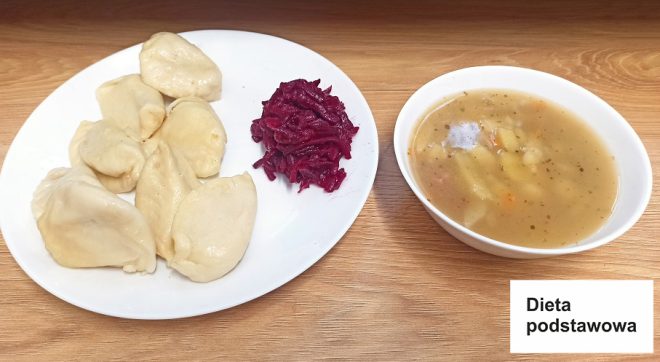 Na tym zdjęciu przedstawiono obiad składający się z: Porcji pierogów, Dodatku z gotowanej lub marynowanej czerwonej kapusty, Miseczki z zupą, przypominającą ziemniaczaną zupę jarzynową. Posiłek jest oznaczony jako „Dieta podstawowa”, co sugeruje, że jest to standardowy posiłek o zrównoważonym składzie, przeznaczony dla osób bez specyficznych ograniczeń dietetycznych