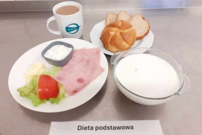 Na zdjęciu widnieje śniadanie oznaczone jako "Dieta podstawowa". Na talerzu znajduje się plaster szynki, kawałek pomidora na liściu sałaty, plasterek masła oraz mała porcja twarogu. Obok leżą kromki chleba oraz bułka. W tle widać filiżankę z napojem (prawdopodobnie kawa z mlekiem) i przezroczystą miseczkę z mlekiem