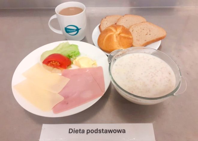 Na zdjęciu widoczny jest zestaw śniadaniowy oznaczony jako "Dieta podstawowa". Na talerzu znajdują się dwa plastry sera żółtego, dwa plastry szynki, liść sałaty, kawałek pomidora i masło. Obok leży bułka oraz dwa kawałki chleba. Na stole znajduje się również miseczka z płatkami owsianymi lub kaszą w mleku oraz kubek z kawą lub herbatą