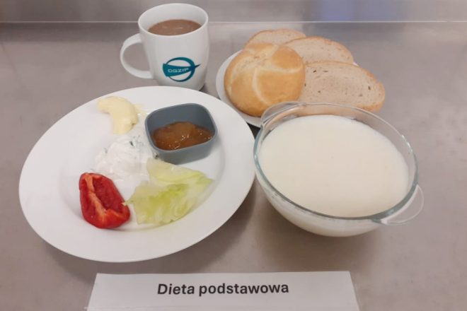 Na zdjęciu widoczne jest śniadanie oznaczone jako "Dieta podstawowa." Na talerzu znajdują się kawałki papryki, liść sałaty, biały ser z ziołami, kostka masła oraz miseczka z dżemem. Obok, na oddzielnym talerzu, znajdują się kromki pieczywa oraz bułka. W tle widoczny jest kubek z napojem oraz miseczka z płynem, prawdopodobnie jogurtem lub mlekiem