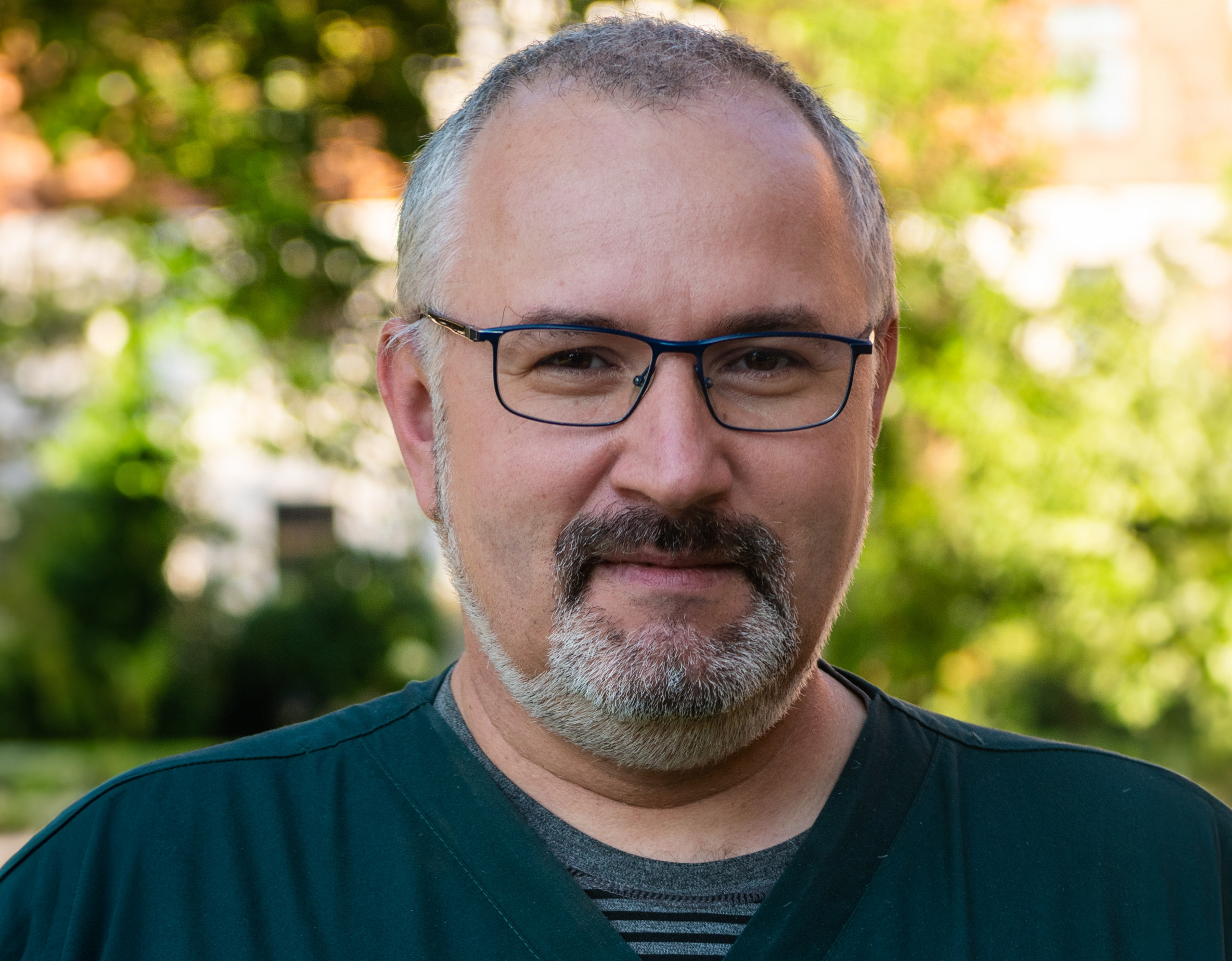 Portret dr n. med. Macieja Chwały, mężczyzny w średnim wieku z brodą, noszącego okulary i zieloną koszulę medyczną, z uśmiechem patrzącego w stronę aparatu. W tle znajduje się rozmyte, naturalne otoczenie