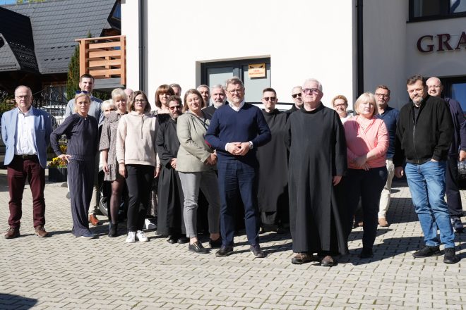 Grupa uczestników konferencji stojących na zewnątrz, uśmiechających się do kamery. W tle widoczny jest budynek, a większość osób stoi w luźnej grupie, niektóre osoby są w habitach zakonnych