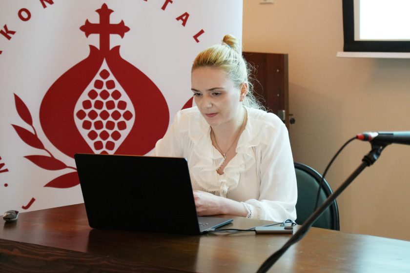 Młoda kobieta siedzi przy laptopie, prowadząc prezentację na konferencji. W tle widoczny jest baner Zakonu Szpitalnego Bonifratrów