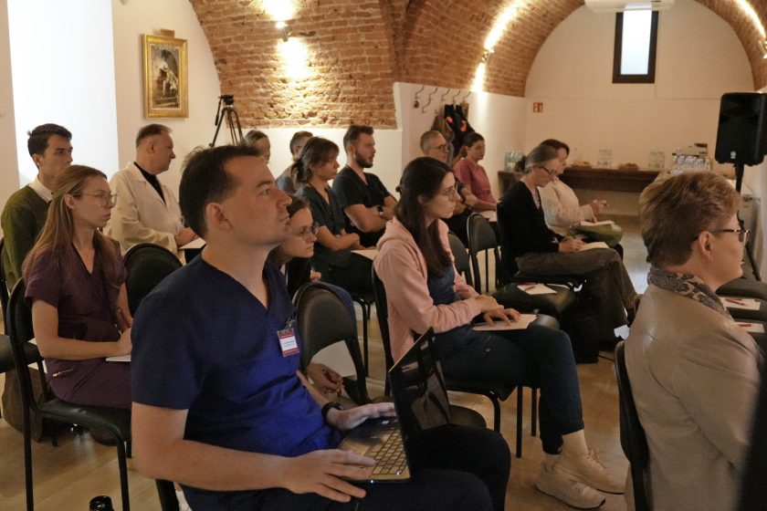 Grupa uczestników konferencji siedzących w sali z ceglaną łukową konstrukcją. Uczestnicy są skupieni, niektórzy z laptopami, inni robią notatki podczas wykładu