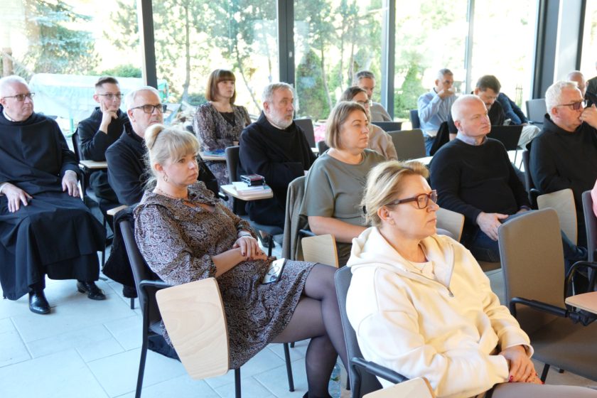 Uczestnicy konferencji siedzący w sali, skupieni na prelekcji. Kilka osób ma na sobie habity, inne są w eleganckich ubraniach, słuchając w ciszy