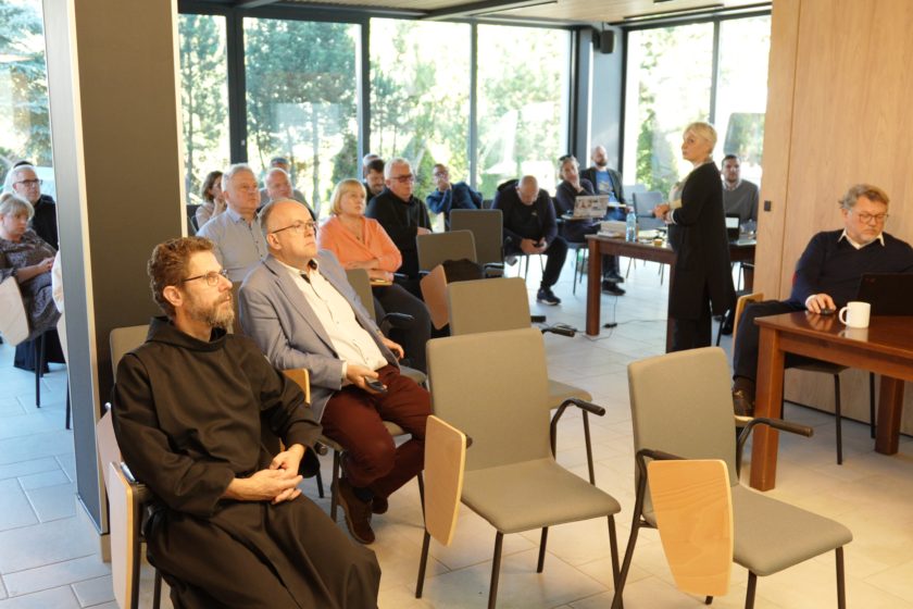 Sala konferencyjna z siedzącymi uczestnikami, w tym zakonnikami i świeckimi osobami, podczas prezentacji. Kobieta prowadząca stoi na przodzie i gestykuluje, a inne osoby w tle słuchają