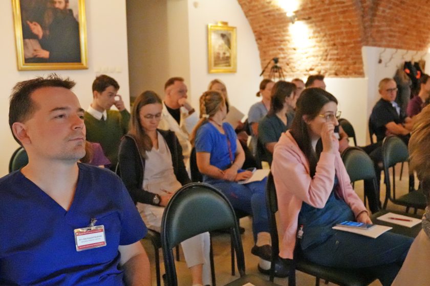 Uczestnicy konferencji siedzą w sali, uważnie słuchając prelekcji. Część osób ubrana w stroje medyczne, widoczne ceglane ściany oraz obrazy na ścianach