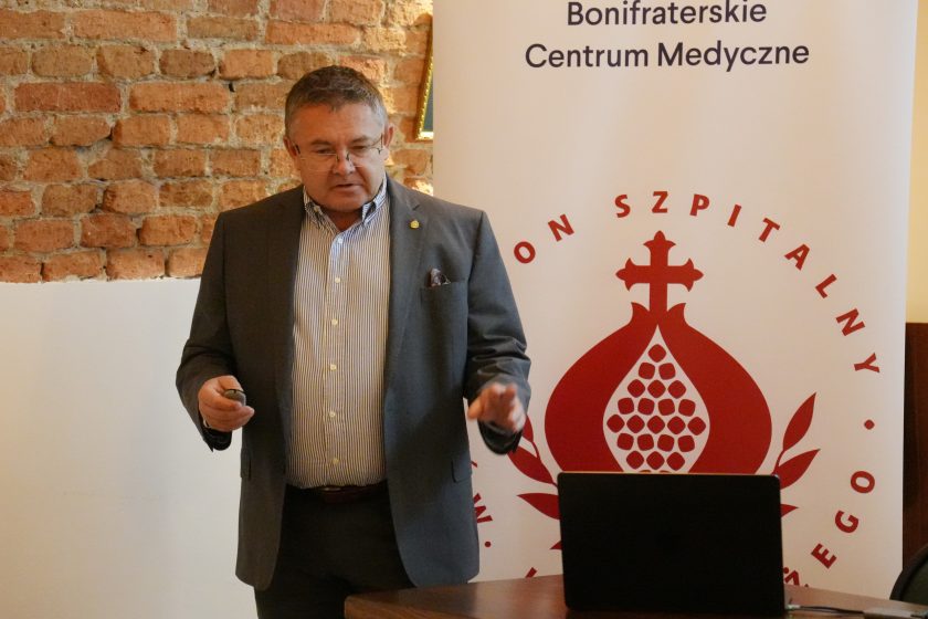 Mężczyzna w garniturze prezentuje swoje wystąpienie przed uczestnikami konferencji, stojąc przy banerze Bonifraterskiego Centrum Medycznego na tle ceglanej ściany