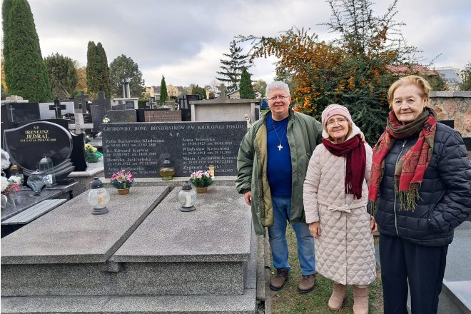 Trzy osoby stoją przy grobie na cmentarzu. Po lewej stronie starszy mężczyzna w zielonej kurtce z krzyżykiem na szyi, pośrodku kobieta w jasnym płaszczu i różowej czapce, a po prawej kobieta w ciemnej kurtce z szalikiem. Za nimi widoczne są inne nagrobki oraz krzew z pomarańczowymi owocami