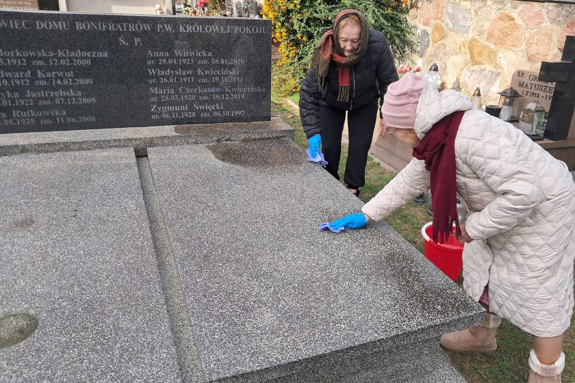 Dwie starsze kobiety czyszczą powierzchnię grobu na cmentarzu. Obie są ubrane w ciepłe kurtki i mają na sobie niebieskie rękawiczki. Na grobie widoczna jest płyta nagrobna z wyrytymi nazwiskami oraz krzew w tle