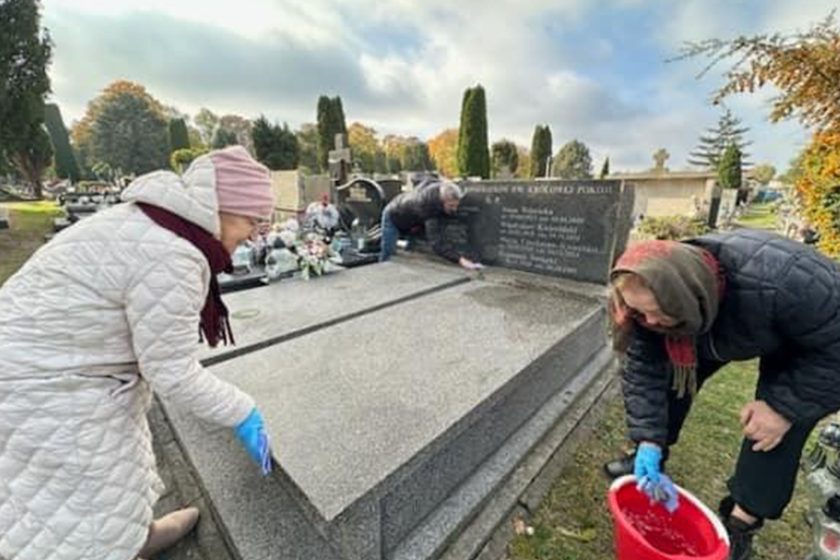 Starsze kobiety sprzątające grób na cmentarzu. Jedna z nich, w jasnym płaszczu i różowej czapce, czyści powierzchnię grobu, trzymając się za krawędź nagrobka, natomiast druga, w ciemnej kurtce, pochyla się z czerwonym wiadrem w dłoni