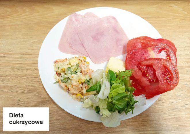 Na zdjęciu widzimy posiłek oznaczony jako „Dieta cukrzycowa”. Na talerzu znajdują się dwa plastry szynki, kilka plastrów pomidora, sałata, kawałek masła oraz mała porcja sałatki warzywnej z kukurydzą. Wszystkie składniki są starannie ułożone na białym talerzu