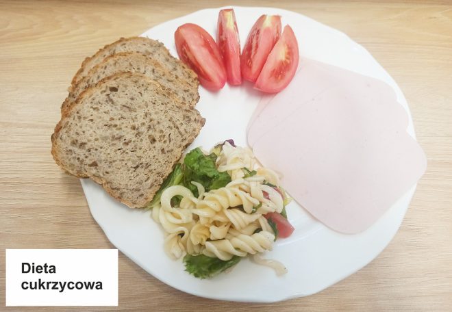 Talerz z kilkoma kromkami pełnoziarnistego chleba, plastrami wędliny drobiowej, kawałkami pomidora oraz sałatką makaronową. Na stole etykieta z napisem „Dieta cukrzycowa”