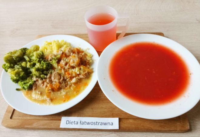 Na drewnianej tacy znajduje się posiłek oznaczony jako "Dieta łatwostrawna". Na białym talerzu znajduje się danie obiadowe, składające się z brokułów, mięsa duszonego z warzywami, ziemniaków oraz sosu. Obok na mniejszym talerzu znajduje się talerz z zupą o czerwonym zabarwieniu, najprawdopodobniej pomidorową. W tle stoi plastikowy kubek z różowym napojem