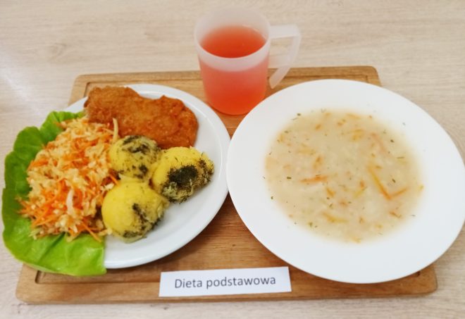 Na zdjęciu przedstawiono posiłek oznaczony jako "Dieta podstawowa". Na tacy znajduje się talerz z trzema porcjami ziemniaków posypanych koperkiem, panierowanym kotletem (najprawdopodobniej schabowym) oraz surówką z marchewki i kapusty, ułożoną na liściu sałaty. Obok talerza znajduje się miska z jasną zupą z kawałkami warzyw, prawdopodobnie jest to zupa jarzynowa. Na tacy jest także przezroczysty kubek z różowym napojem, który może być kompotem lub herbatą owocową. Całość ułożona jest na drewnianej tacy