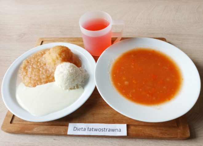 Na drewnianej tacy znajduje się posiłek oznaczony jako "Dieta łatwostrawna". Na talerzu z lewej strony znajduje się ryż, puree ziemniaczane i przecier jabłkowy, polane białym sosem. Na drugim talerzu widoczna jest zupa, najprawdopodobniej pomidorowa z drobnymi kawałkami warzyw. Obok znajduje się kubek z różowym napojem, prawdopodobnie kompotem lub herbatą owocową