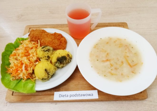 Zestaw obiadowy zawiera panierowany kotlet, porcję surówki z marchwi i kapusty, oraz trzy kulki ziemniaczanego purée posypane koperkiem, podane na białym talerzu z dodatkiem sałaty. Obok znajduje się miseczka z zupą o jasnym kolorze z dodatkami warzyw. Całość jest ułożona na drewnianej desce, a w rogu widnieje napis „Dieta podstawowa”. Obok talerzy stoi kubek z różowym napojem