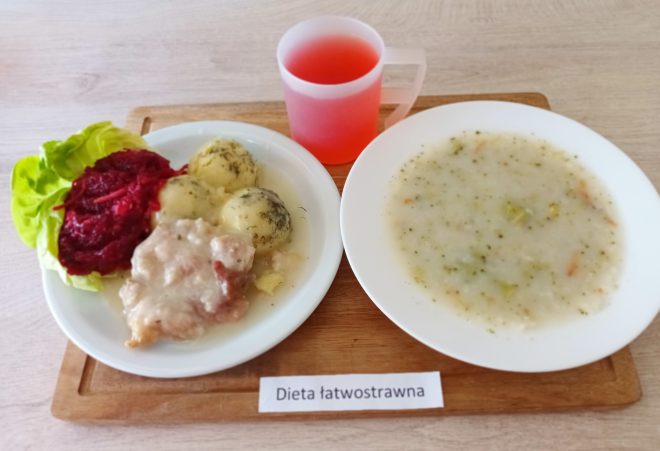 Zdjęcie przedstawia posiłek oznaczony jako „dieta łatwostrawna”. Na drewnianej tacce znajdują się: talerz z daniem głównym, na którym są gotowane ziemniaki posypane koperkiem, sos z kawałkami mięsa, a także buraczki na liściu sałaty, miseczka z zupą jarzynową o delikatnej konsystencji, z kawałkami warzyw, kubek z czerwoną herbatą lub kompotem. Danie jest serwowane na białych naczyniach