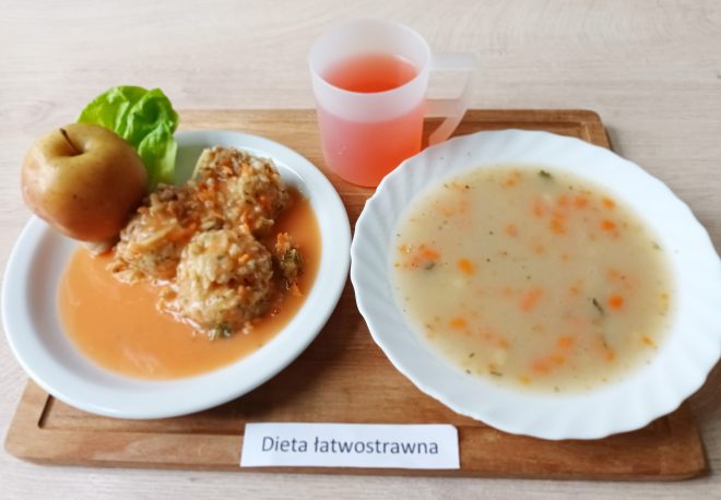 Na drewnianej tacy przedstawiono zestaw obiadowy oznaczony jako "Dieta łatwostrawna". W jego skład wchodzi zupa podana na białym, falistym talerzu – kremowa z drobnymi kawałkami marchewki i pietruszki, przyprawiona ziołami, prawdopodobnie z dodatkiem ziemniaków. Obok zupy na małym białym talerzyku znajdują się trzy pulpety w sosie warzywnym, dekorowane liściem sałaty. Na talerzu znajduje się również całe jabłko. Do zestawu dołączono napój o różowawym odcieniu, podany w przezroczystym plastikowym kubku z uchwytem. Całość prezentuje się skromnie, apetycznie i lekkostrawnie, odpowiednia dla osób potrzebujących łagodnej, łatwostrawnej diety