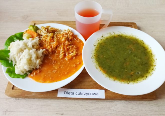 Na zdjęciu znajduje się obiad oznaczony karteczką z napisem „Dieta cukrzycowa”. Zestaw składa się z talerza z makaronem w sosie pomidorowym oraz surówki z kapusty i warzywnych dodatków na liściu sałaty. Obok znajduje się talerz z zieloną zupą. Zestaw uzupełnia kubek z czerwonawym napojem