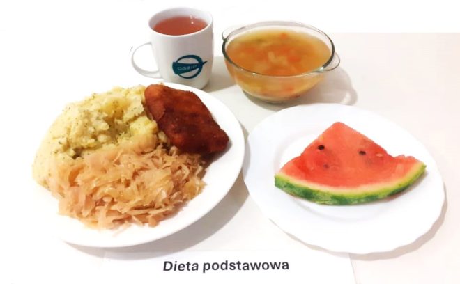 Na zdjęciu widzimy obiad w ramach diety podstawowej. Na talerzu znajdują się ziemniaki puree z koperkiem, kapusta kiszona oraz panierowany kotlet. Na osobnym talerzyku położono kawałek arbuza. Obok posiłku znajduje się miska z zupą jarzynową oraz kubek z ciepłym napojem, prawdopodobnie herbatą