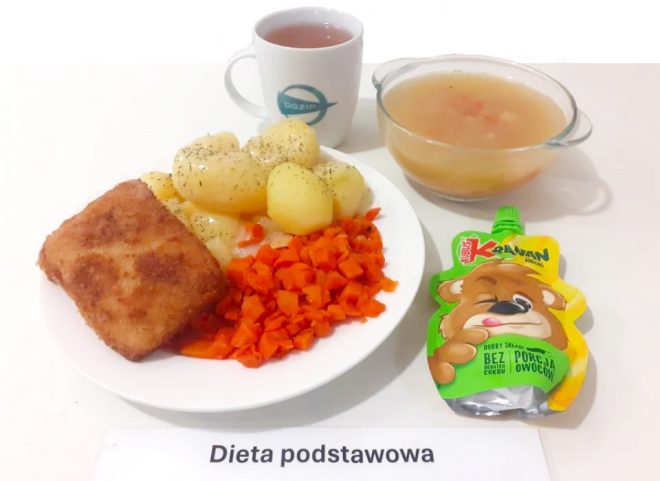 Na zdjęciu przedstawiono posiłek oznaczony jako "Dieta podstawowa". Na talerzu znajduje się panierowany kotlet, ugotowane ziemniaki posypane ziołami oraz gotowana marchewka. Obok widoczna jest miseczka z klarowną zupą oraz kubek z herbatą. Po prawej stronie znajduje się owocowy mus w opakowaniu z wizerunkiem misia