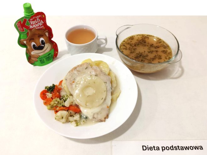 Na zdjęciu przedstawiony jest obiad oznaczony jako "Dieta podstawowa". Na talerzu znajduje się kawałek mięsa z kurczaka w sosie z plasterkiem ananasa, porcja gotowanych ziemniaków oraz mieszanka warzyw gotowanych (brokuły, kalafior, marchewka). Obok na stole widoczna jest miseczka z zupą i kubek kompotu. W tle stoi saszetka z musem owocowym „Truskawka”. Na stole umieszczona jest karteczka z napisem "Dieta podstawowa"