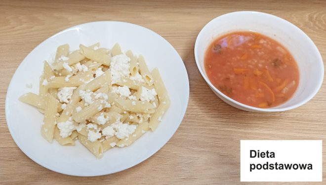 Na zdjęciu widzimy posiłek oznaczony jako "Dieta podstawowa". Na talerzu znajduje się makaron penne z białym serem. Obok talerza stoi miska z zupą pomidorową, w której znajdują się kawałki warzyw, w tym marchewka i ryż. Całość jest podana na drewnianym blacie