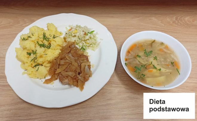 Obiad oznaczony jako „Dieta podstawowa”. Na białym talerzu znajduje się porcja tłuczonych ziemniaków posypanych koperkiem, duszona cebula z mięsem oraz surówka z białej kapusty i warzyw. Obok na talerzu znajduje się miseczka z zupą jarzynową, w której widać kawałki marchewki i pietruszki, udekorowaną natką pietruszki