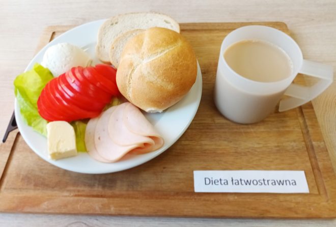 Na drewnianej tacy znajduje się posiłek oznaczony jako "Dieta łatwostrawna". Na białym talerzu ułożono śniadanie, składające się z kromki chleba, bułki, plasterków wędliny drobiowej, masła, plasterków pomidora, liścia sałaty oraz jajka na twardo. Obok stoi plastikowy kubek z jasnym napojem, najprawdopodobniej herbatą z mlekiem