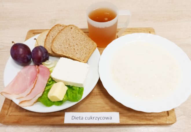 Na zdjęciu przedstawiono posiłek oznaczony jako "Dieta cukrzycowa". Na tacy znajduje się kilka składników: dwie kromki chleba pełnoziarnistego, dwie śliwki, trzy plasterki wędliny, porcja białego sera, liść sałaty z kawałkiem masła oraz filiżanka z napojem (najprawdopodobniej herbatą). Obok talerza z tymi produktami znajduje się talerz z płynną potrawą, która może być zupą mleczną. Całość posiłku jest ułożona na drewnianej tacy