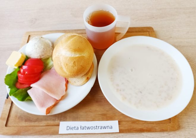 Na zdjęciu przedstawiono posiłek oznaczony jako "Dieta łatwostrawna". Na talerzu znajdują się dwa plasterki szynki, kilka plastrów pomidora, liść sałaty, niewielka porcja masła, bułka oraz kulka twarogu. Obok na drewnianej desce stoi filiżanka z herbatą oraz miska z owsianką lub kleikiem mlecznym. Cały posiłek znajduje się na jasnym stole.