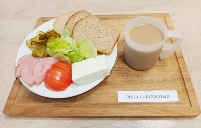 Na tacy znajduje się śniadanie oznaczone jako „Dieta cukrzycowa”. Na białym talerzu leżą trzy kromki jasnego chleba, kilka plastrów szynki, kawałek białego sera, plasterki pomidora, liście sałaty oraz plasterki ogórka kiszonego. Obok talerza stoi plastikowy kubek z jasnobrązowym napojem, prawdopodobnie kawą z mlekiem.