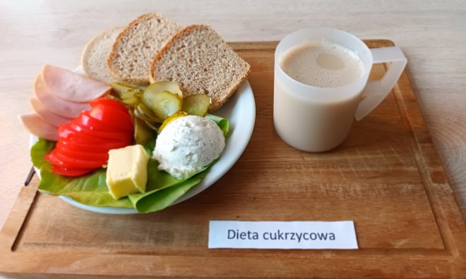 Na drewnianej tacy znajduje się posiłek oznaczony jako "Dieta cukrzycowa". Na talerzu widoczne są trzy kromki chleba pełnoziarnistego, plasterki szynki, pokrojony pomidor, plasterki kiszonego ogórka, porcja masła oraz kulka serka twarogowego na liściu sałaty. Obok znajduje się kubek z białym napojem, prawdopodobnie mlekiem lub napojem mlecznym