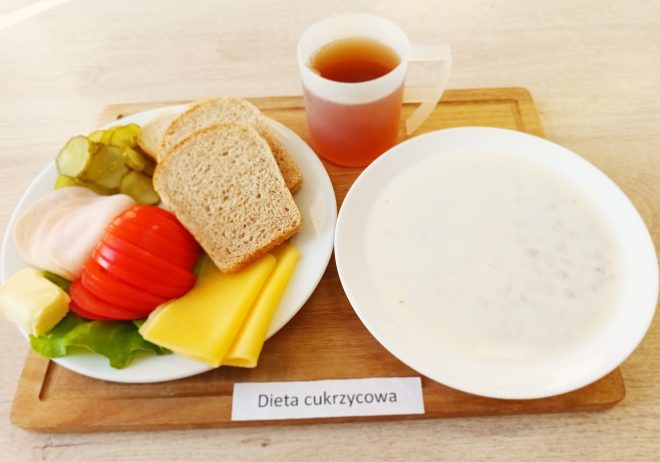 Na zdjęciu przedstawione jest śniadanie oznaczone jako "Dieta cukrzycowa". Na drewnianej tacce znajduje się talerz z kilkoma kromkami chleba pełnoziarnistego, plasterkami świeżego pomidora, ogórków kiszonych, sałatą, plasterkami żółtego sera oraz wędliną. Obok talerza stoi kubek z ciemnym napojem, prawdopodobnie herbatą. Na tacy znajduje się także głęboki talerz z białą zupą mleczną z widocznymi płatkami lub drobnymi zbożami