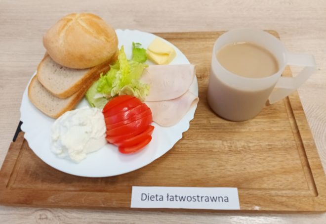 Na drewnianej tacy znajduje się śniadanie oznaczone jako "Dieta łatwostrawna". Na białym talerzu ułożono bułkę, dwa kawałki chleba tostowego, kawałek masła, plasterki pomidora, dwa liście sałaty, plasterki wędliny oraz porcję serka śmietankowego. Obok talerza stoi przezroczysty kubek z jasnobrązowym napojem, prawdopodobnie herbatą z mlekiem