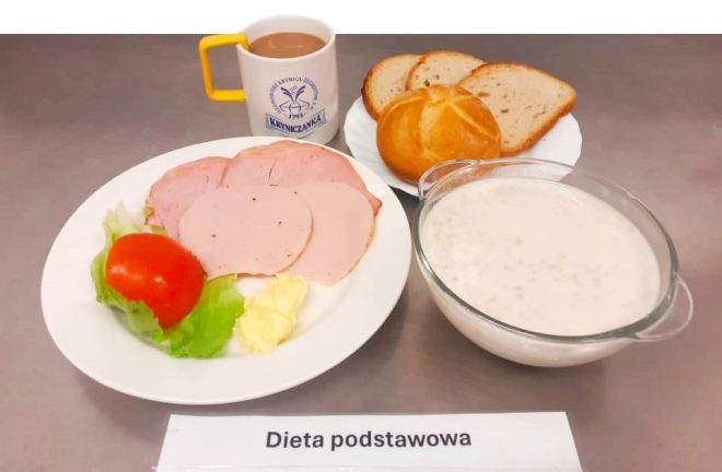 Na zdjęciu znajduje się śniadanie oznaczone jako „Dieta podstawowa”. Na talerzu leży kilka plastrów wędliny z dodatkiem sałaty, plasterka pomidora oraz masła. Obok znajduje się miseczka z białą potrawą (prawdopodobnie owsianką lub zupą mleczną), filiżanka kawy oraz talerzyk z kromkami chleba i bułką