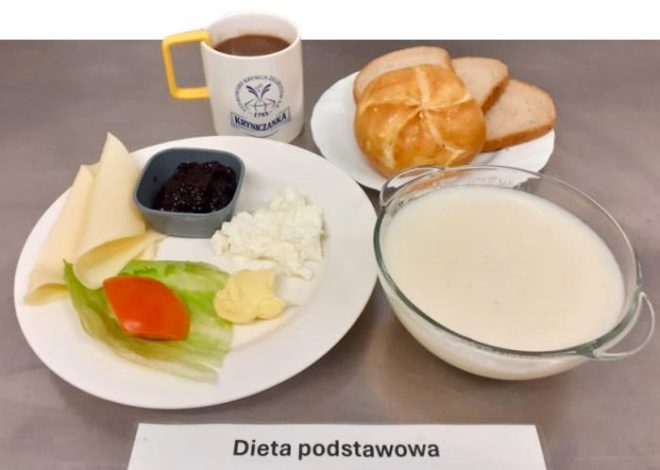 Na zdjęciu przedstawiono posiłek oznaczony jako "Dieta podstawowa". Na talerzu znajdują się plasterek sera, odrobina twarogu, plasterek pomidora z liściem sałaty oraz masło. Obok umieszczono bułkę oraz kromki chleba. W miseczce widoczny jest biały płyn (prawdopodobnie mleko lub kasza manna). Z boku znajduje się kubek z ciepłym napojem oraz dżem w małej miseczce