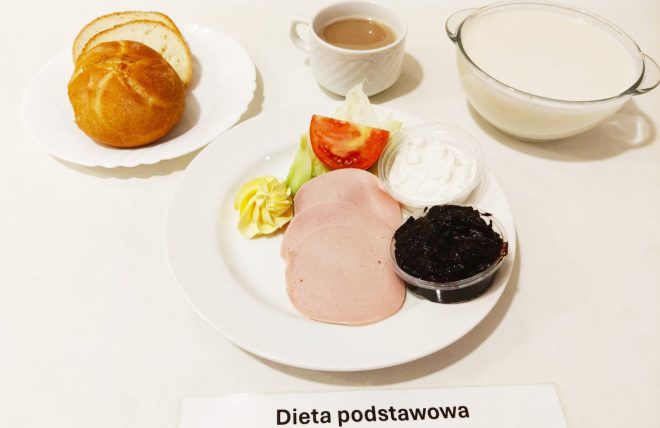 Posiłek oznaczony jako "Dieta podstawowa". Na talerzu znajduje się kilka plastrów szynki, kawałek sałaty, plaster pomidora, mała porcja białego twarożku oraz porcja dżemu. Obok umieszczono dekoracyjnie porcję masła. Na osobnym talerzyku leżą bułka i kromka chleba. Posiłek uzupełnia filiżanka kawy z mlekiem oraz miska z zupą mleczną