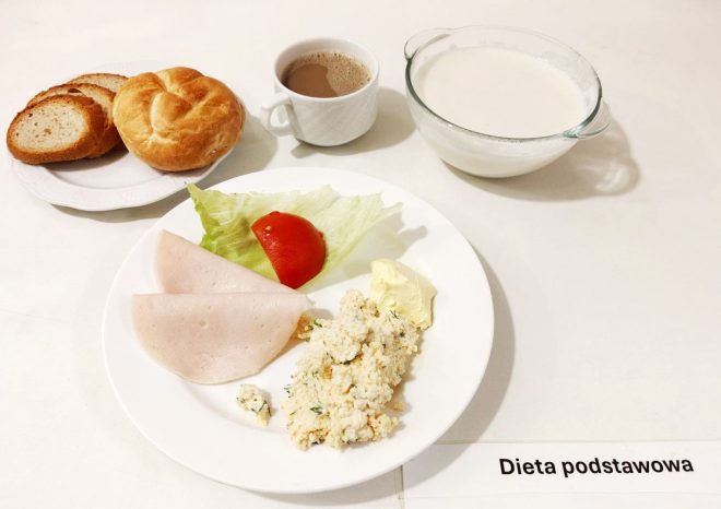 Śniadanie oznaczone jako „Dieta podstawowa”. Na białym talerzu znajduje się sałata lodowa, plasterek pomidora, dwa plastry drobiowej wędliny, porcja twarożku z koperkiem oraz kawałek masła. Obok na talerzu leży bułka oraz trzy kromki chleba (jasnego i razowego). Na tacy znajduje się filiżanka z napojem (prawdopodobnie kawa z mlekiem) oraz miseczka z mlekiem, najprawdopodobniej przeznaczona do płatków lub kaszy