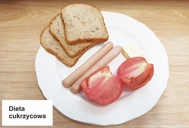 Na białym talerzu znajduje się śniadanie oznaczone jako „Dieta cukrzycowa”. Na talerzu ułożono trzy kromki chleba, dwie parówki drobiowe, kawałek masła oraz połówkę pomidora