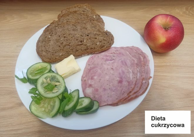 Na talerzu znajduje się śniadanie oznaczone jako "Dieta cukrzycowa". Składa się z dwóch kromek pełnoziarnistego chleba, kilku plasterków wędliny, kawałka masła oraz kilku plasterków świeżego ogórka z cebulką dymką. Na talerzu obok leży jabłko, które jest częścią zestawu śniadaniowego. Na stole widoczna jest karteczka z napisem "Dieta cukrzycowa"