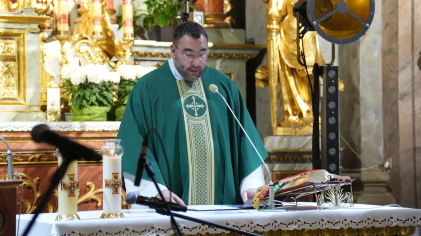 Ksiądz ubrany w zieloną szatę liturgiczną stoi przy ołtarzu, odczytując modlitwy. W tle złocone elementy ołtarza oraz świece symbolicznie oświetlające miejsce