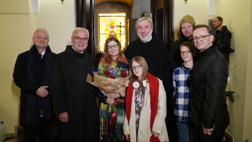 Grupa osób pozuje do zdjęcia w wejściu do kościoła. W centralnej części kobieta z bukietem kwiatów, otoczona przez duchownych i dwójkę dzieci. W tle witraż z motywami religijnymi