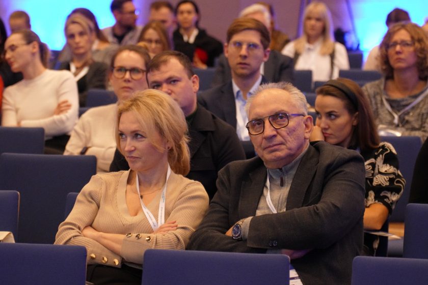 Sala konferencyjna wypełniona uczestnikami w eleganckich strojach. Na pierwszym planie mężczyzna w ciemnym garniturze i kobieta w jasnym swetrze siedzą z założonymi rękami, obserwując z uwagą przebieg wydarzenia. W tle widoczni inni uczestnicy skupieni na wykładzie, z niebieskim oświetleniem nadającym atmosferę profesjonalizmu