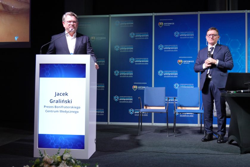 Prelegent w ciemnym garniturze stoi przy mównicy z napisem "Jacek Graliński", trzyma mikrofon. W tle widoczna jest scena z profesjonalnym oświetleniem i tablicami konferencyjnymi w kolorze niebieskim