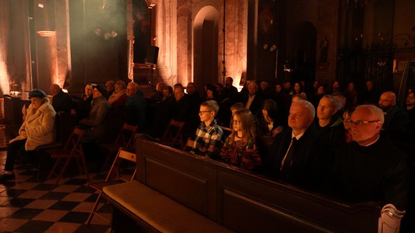 Publiczność zgromadzona w kościele, siedząca w ławkach i na krzesłach. Atmosfera jest nastrojowa, z delikatnym oświetleniem akcentującym detale wnętrza