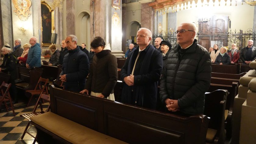 Wierni stojący w ławkach kościelnych podczas mszy. Na pierwszym planie mężczyzna w puchowej kurtce oraz kobieta w ciemnym płaszczu, trzymający dłonie złożone w geście modlitwy