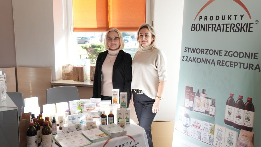 Dwie kobiety stojące przy stoisku promującym „Produkty Bonifraterskie”. Na stole przed nimi znajdują się różnorodne produkty medyczne i zdrowotne w opakowaniach. W tle widoczny jest baner z logo „Produkty Bonifraterskie” oraz informacją o ich recepturze zakonnej