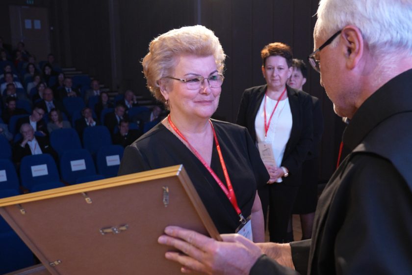 Elegancko ubrana kobieta w czarnej sukience z przypiętą plakietką odbiera dyplom z rąk zakonnika. W tle widać zapełnioną widownię oraz osoby oczekujące na swoją kolej do odebrania wyróżnień